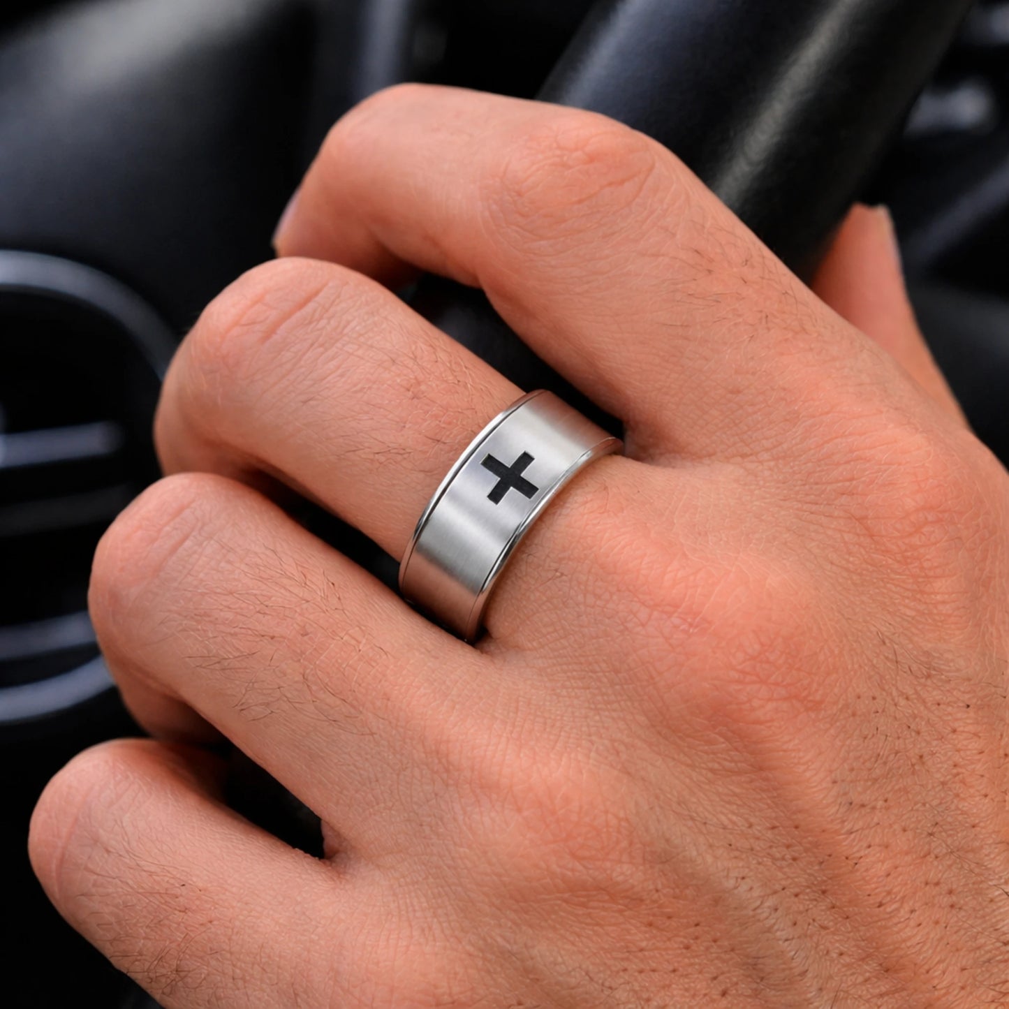 Hand wearing a silver ring with a black cross symbol, blurred car interior background