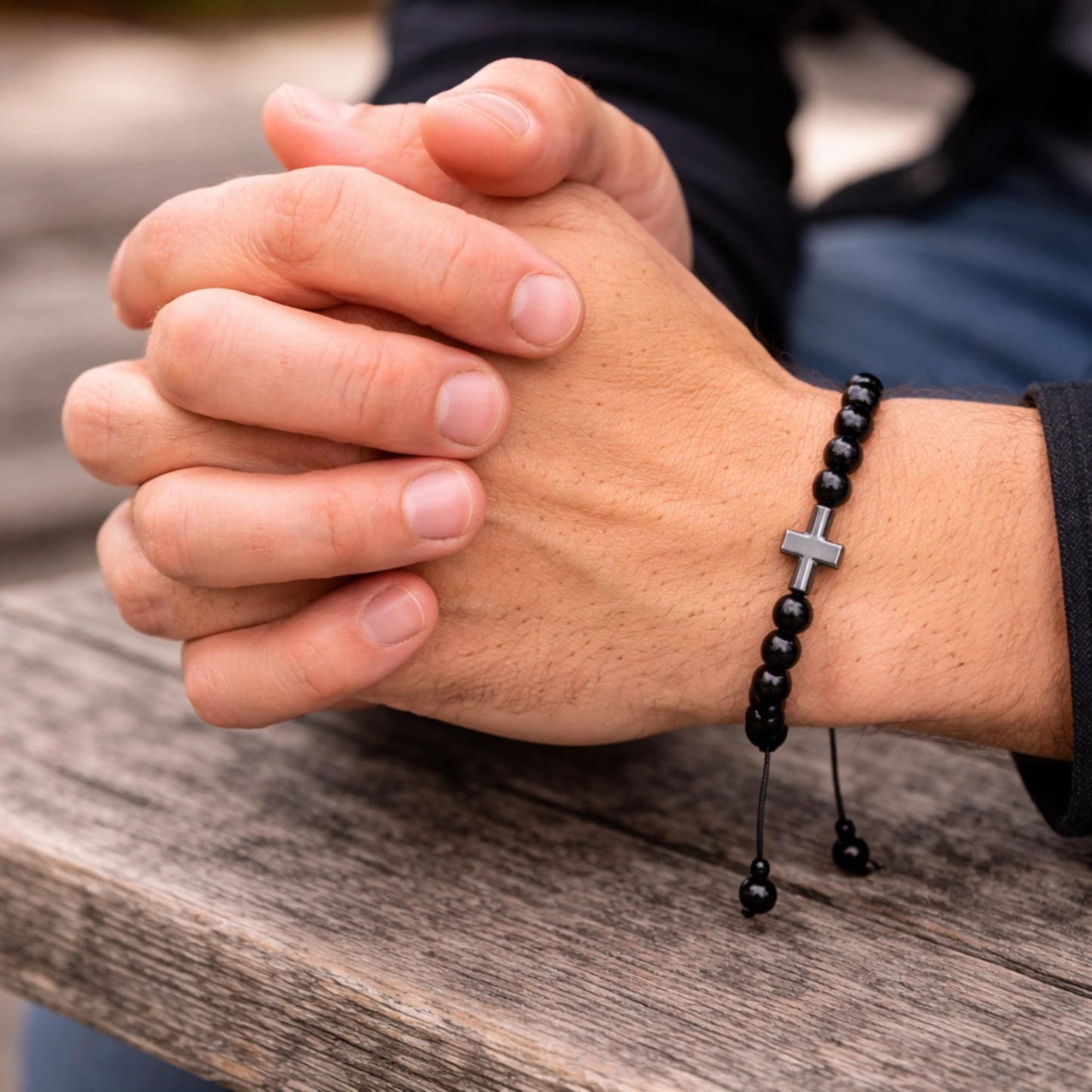 Close-up of hands clasped together with a black beaded bracelet featuring a cross on a wooden surface.